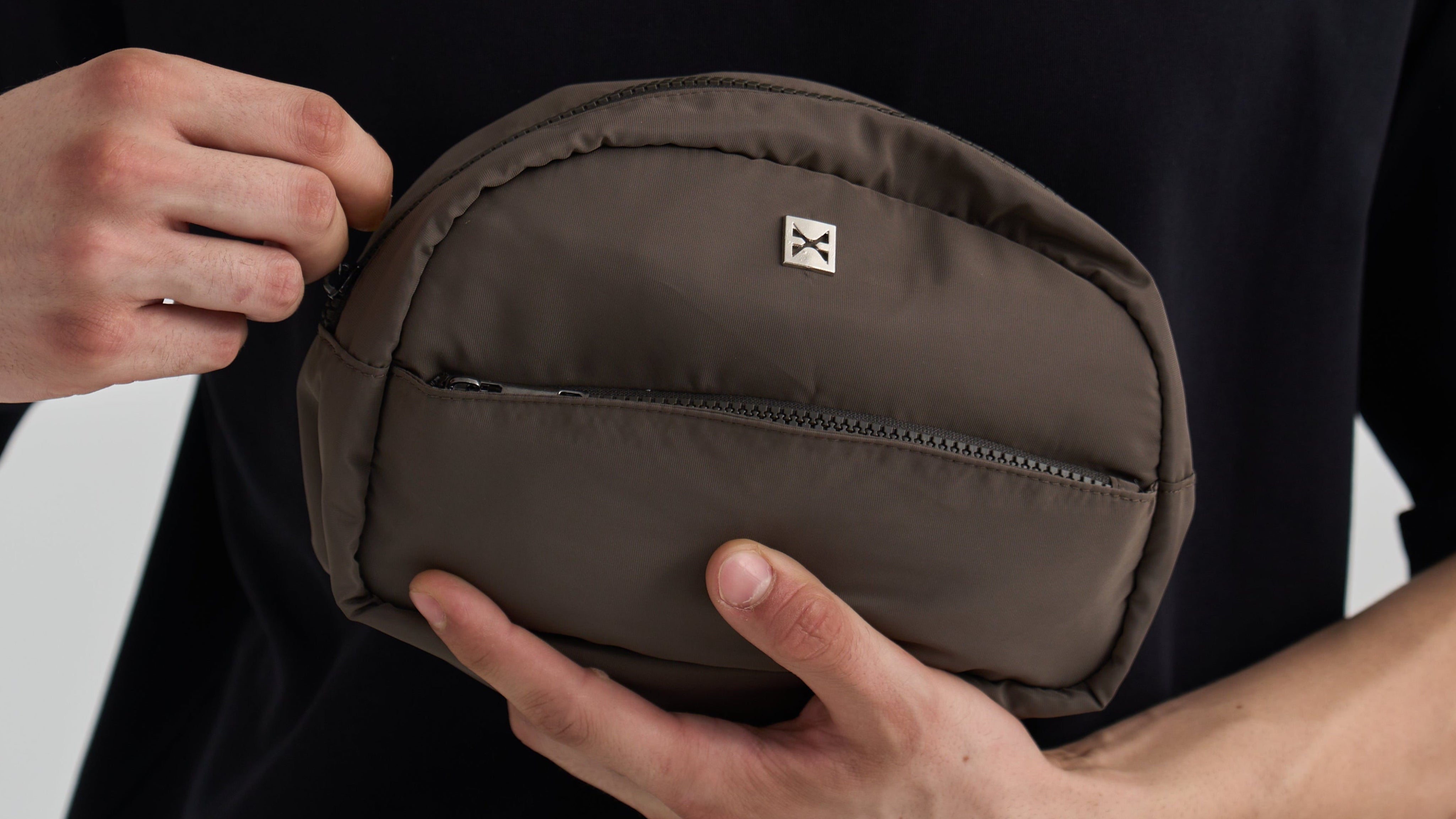 A person in a black shirt holds a small oval brown nylon pouch with a front zipper and a small white square logo patch. The person's hands and torso are visible against a light gray background.